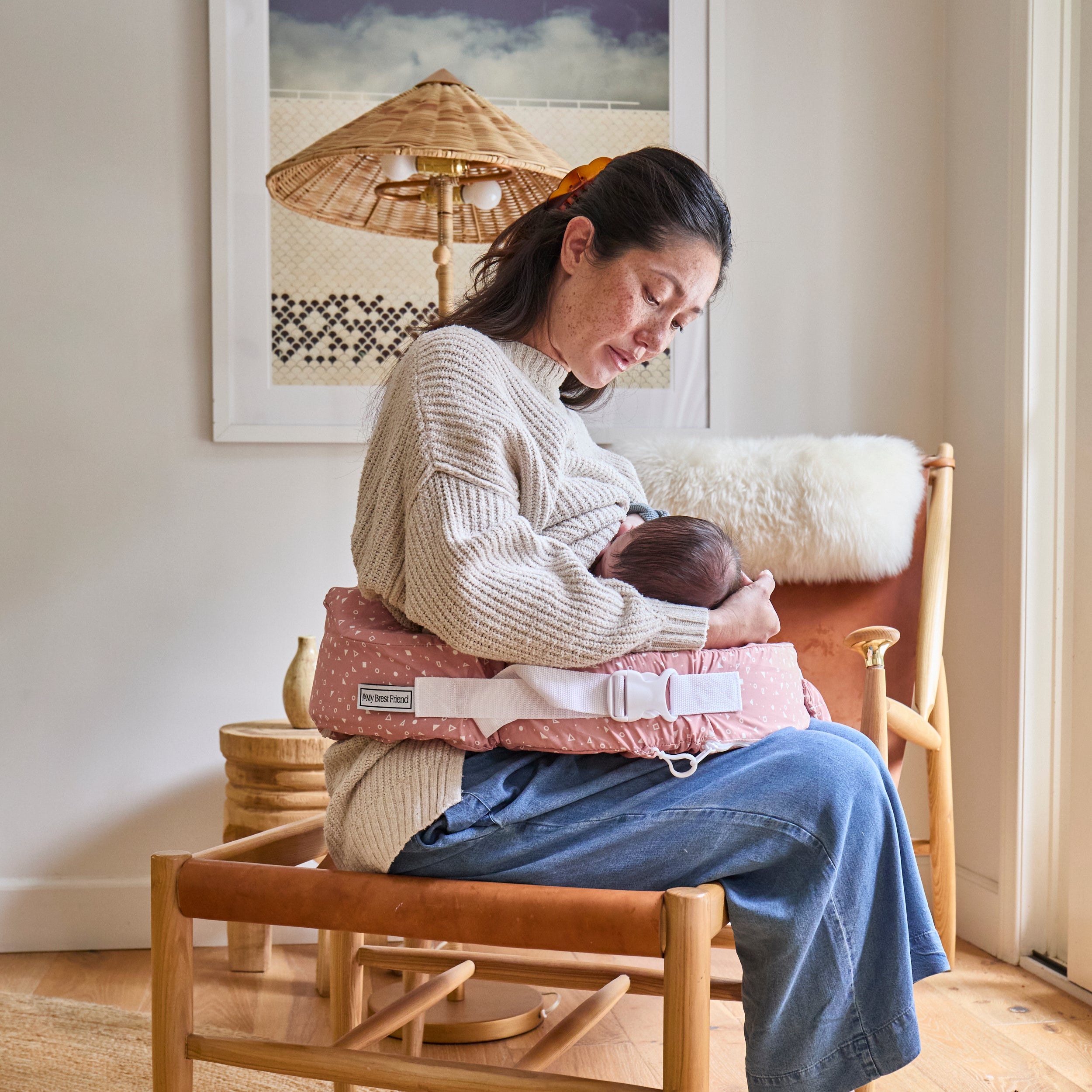 Woman sitting on a chair holding a baby in a sling, with a framed picture on the wall behind her.