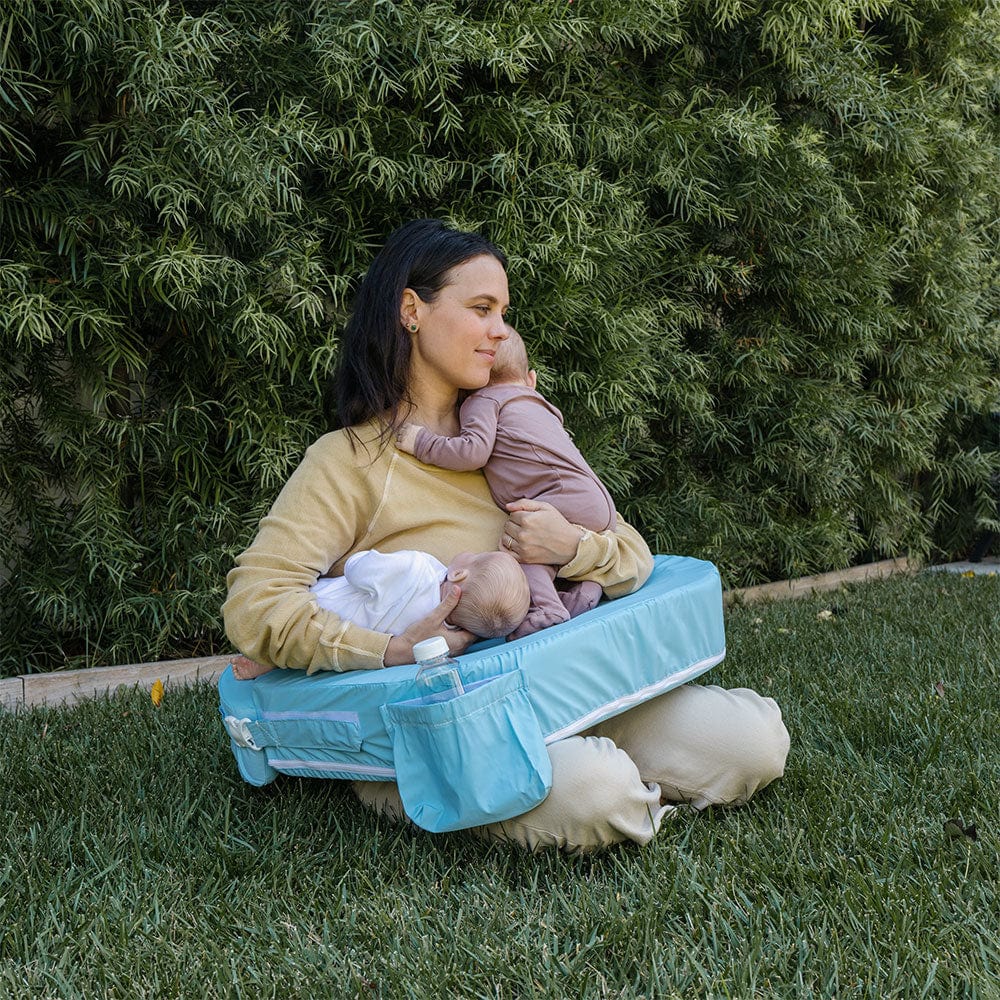Woman sitting on a portable changing pad with two babies outdoors.