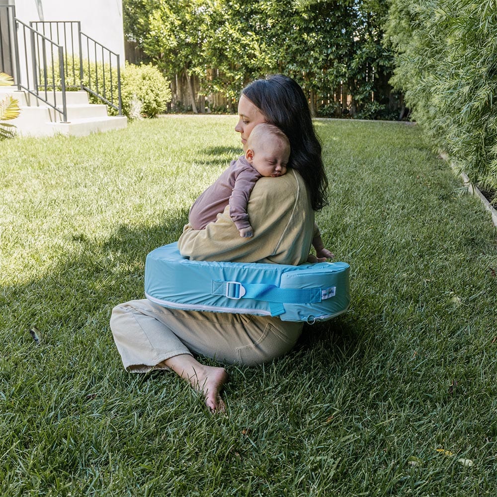 Woman sitting on grass holding a baby in a blue carrier