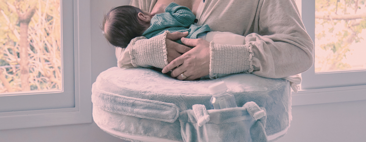 Person holding a baby on a cushioned ottoman in a softly lit room.