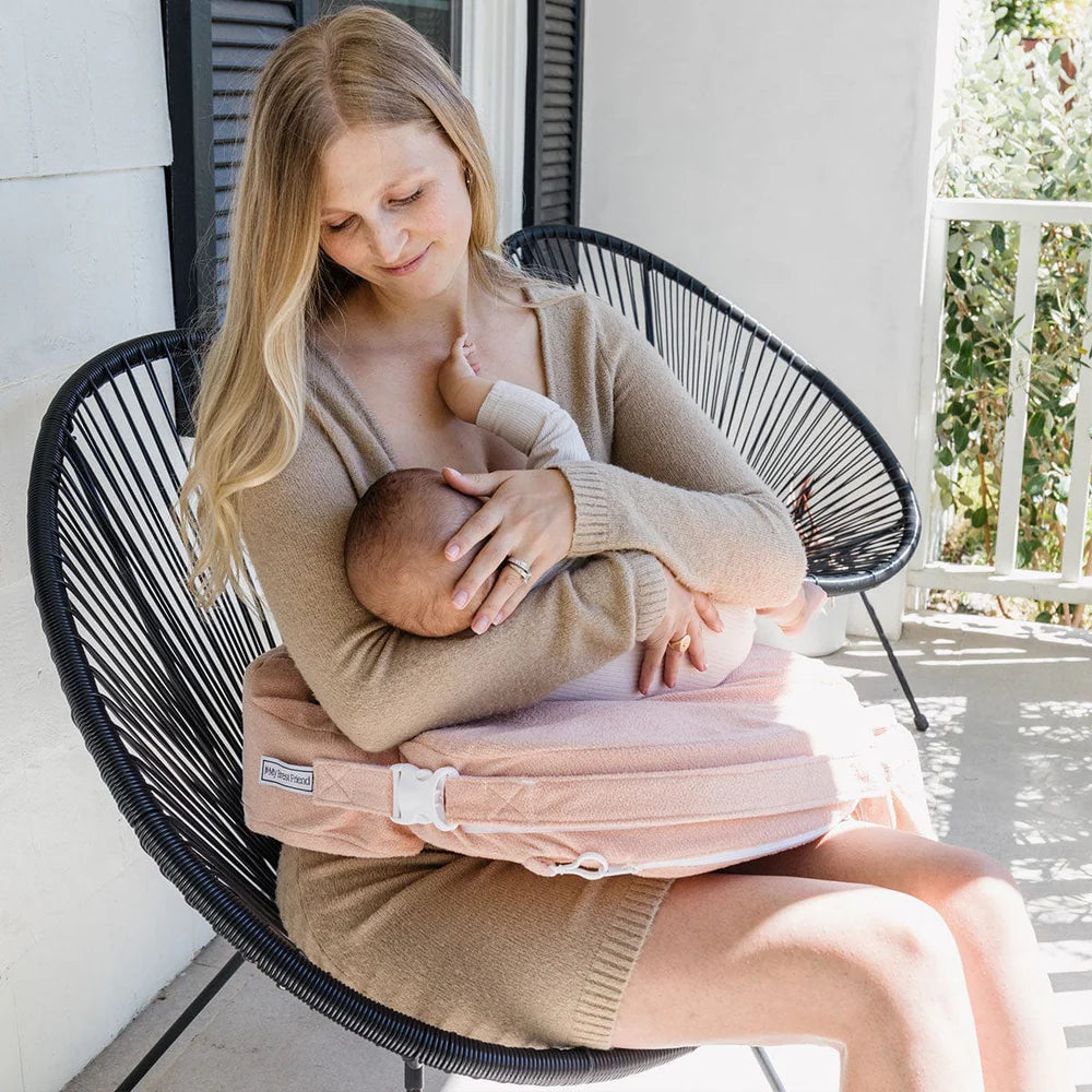 A mother breastfeeds her baby on a chair, using a pink nursing pillow. She wears a beige sweater and smiles down with love and care, in a serene outdoor setting.