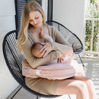 A mother breastfeeds her baby on a chair, using a pink nursing pillow. She wears a beige sweater and smiles down with love and care, in a serene outdoor setting.