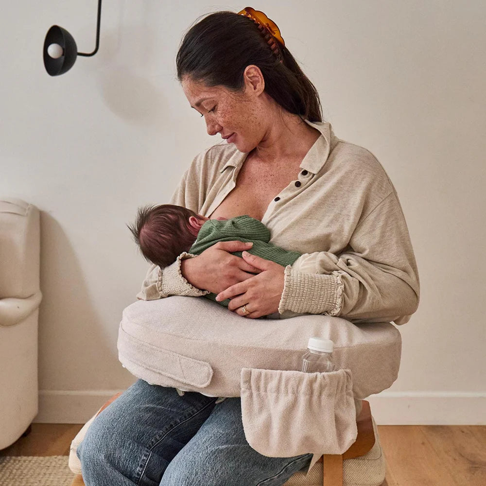 A woman breastfeeds her baby using a beige nursing pillow with a pocket for a bottle. She sits in a cozy room, smiling softly and looking down.
