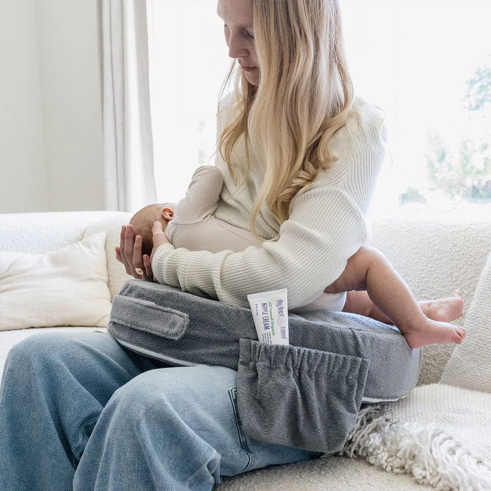 A woman with long blonde hair is breastfeeding a baby on a couch. She's using a gray nursing pillow, wearing a cream sweater and jeans. The atmosphere is calm and nurturing.