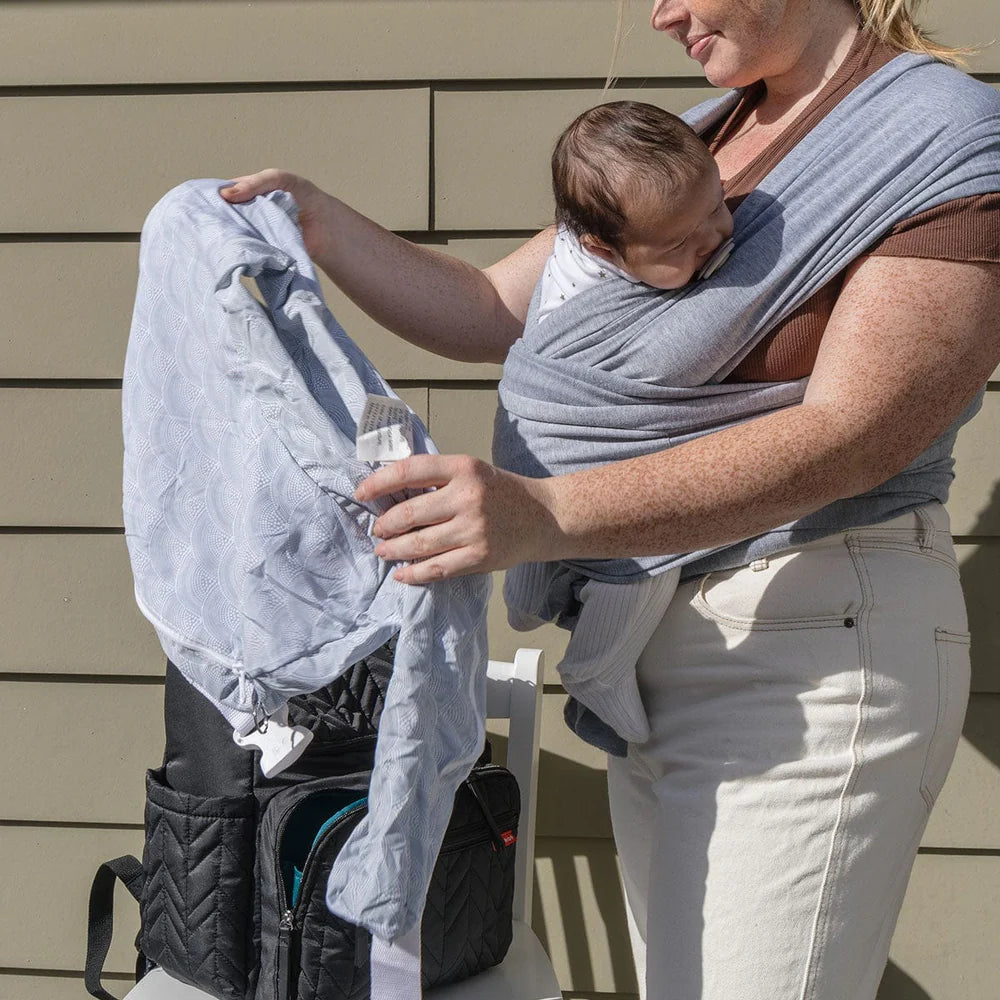 Woman holding a baby in a sling and opening a backpack outdoors.