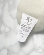 Plastic tube of Lanolin Creme by LANACare sits on beige fabric. The label features a circular sheep logo. The scene conveys a sense of softness.