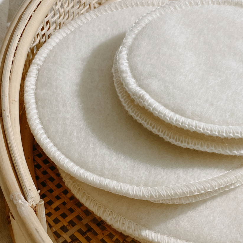 A neatly stacked set of white LANACare merino wool nursing pads resting on a woven basket — soft, breathable, and naturally balanced for your feeding setup.
