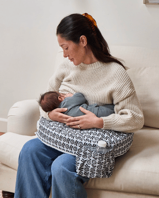Woman breastfeeding a baby using a nursing cover on a white couch.