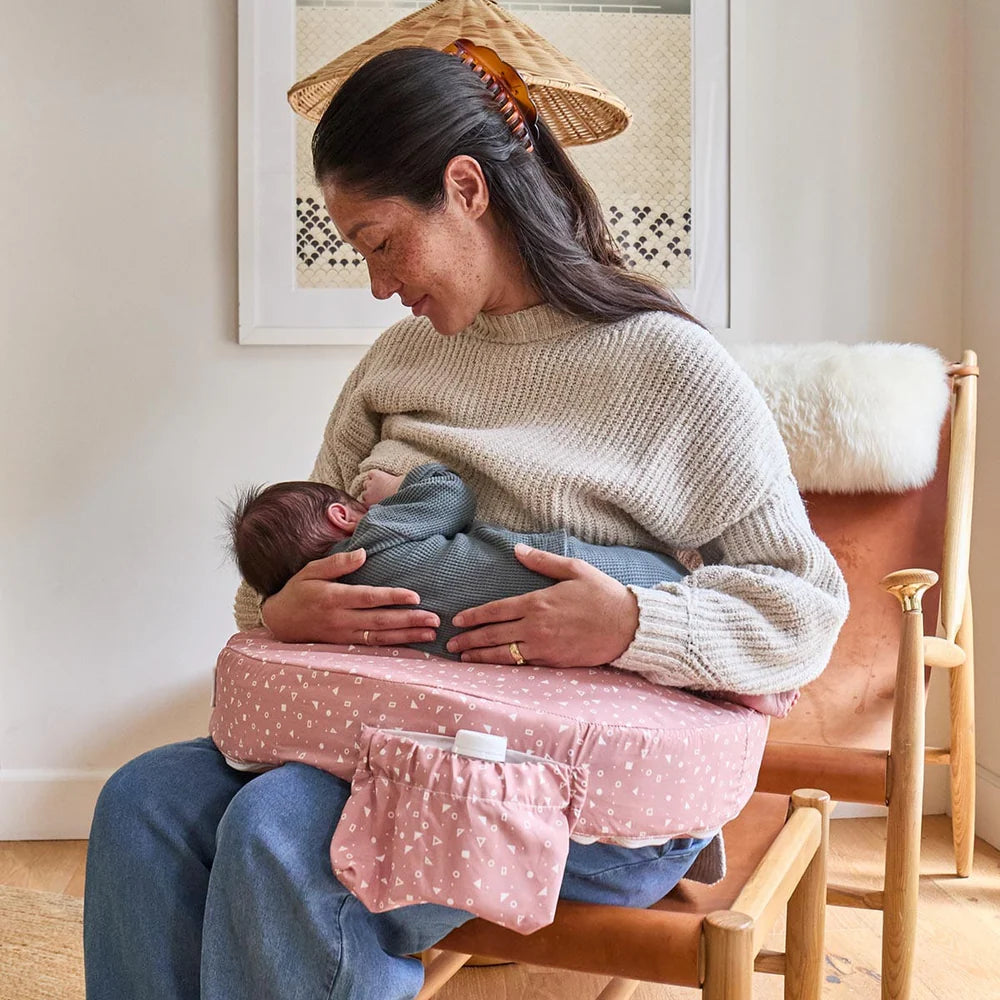 Woman breastfeeding a baby using a pink nursing pillow in a cozy room.