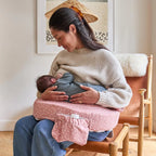 Woman breastfeeding a baby using a pink nursing pillow in a cozy room.