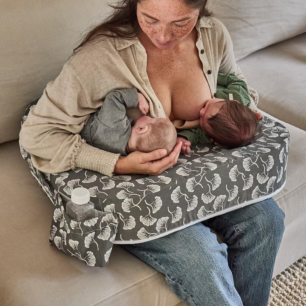 Woman breastfeeding two babies on a patterned cushion in a cozy living room.