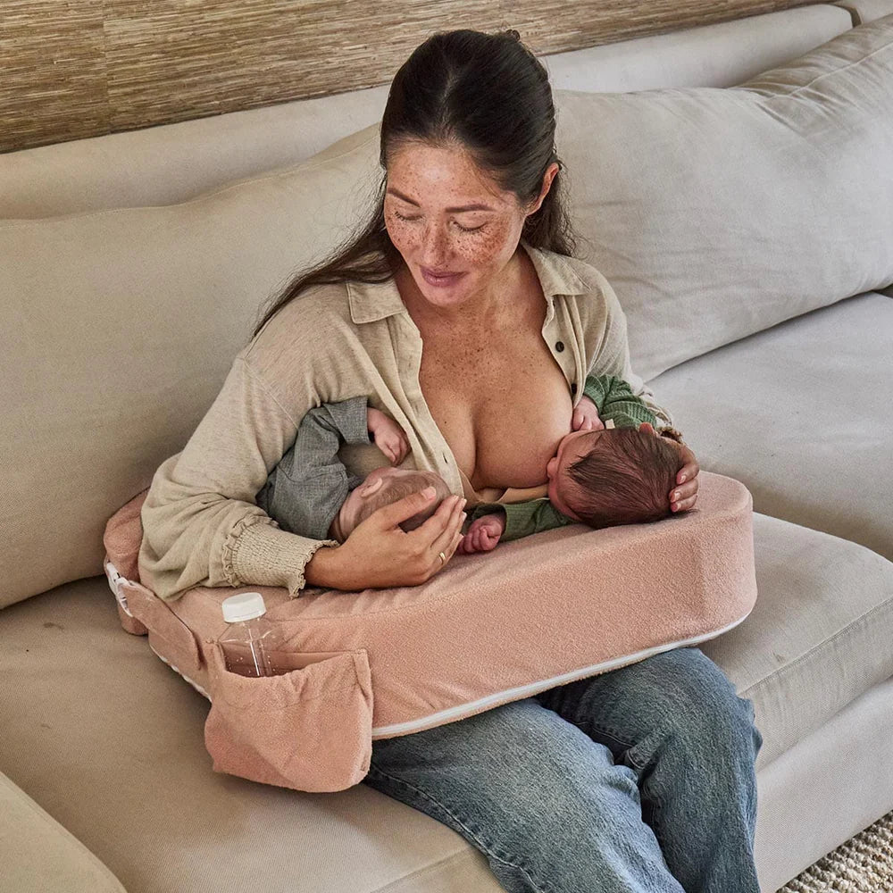 Woman breastfeeding twins using a pink哺乳枕 on a couch.