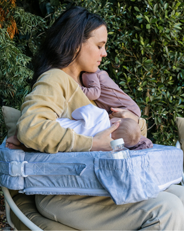 Woman sitting outdoors with a baby in a carrier, holding a bottle.
